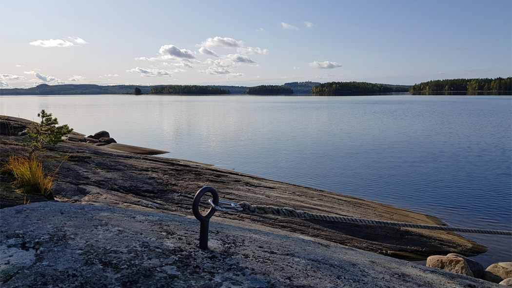 En lugn sjö - skärgårdslandskap i tidigt kvällsljus. I förgrunden finns en lätt sluttande, låg klippa med ett fäste för båtar.