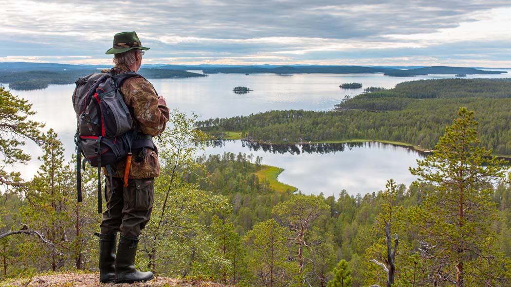 En vandrare med ryggsäck står på en utsiktsplats upp på en hög backe. Ett grönskande somrigt sjö- och skogslandskap kan ses från backen. Himlen är molnig.