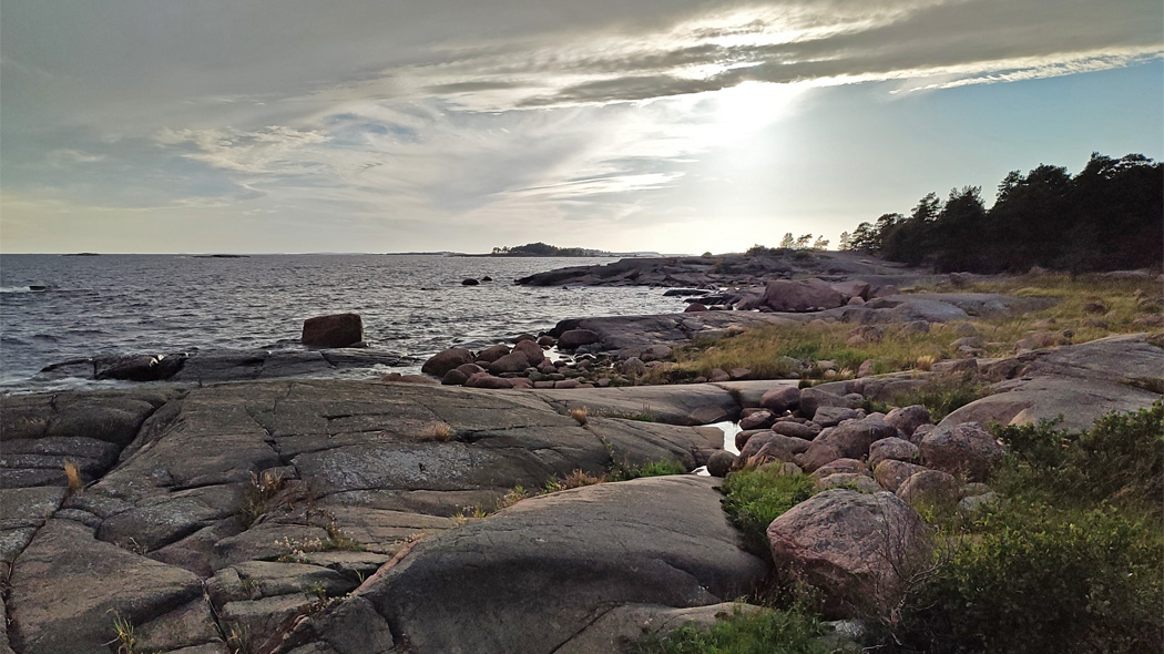 Skärgårdslandskap. I förgrunden släta klippor. I bakgrunden skog, stenig strand och horisont. Solen lyser bakom molnen.