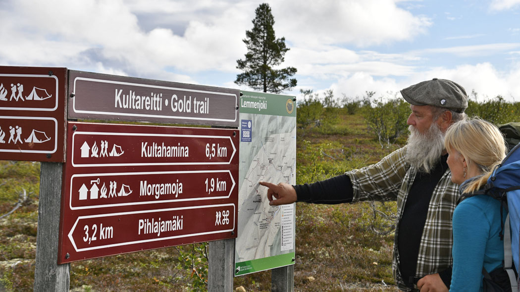 Det ligger en lång öppen båt på floden där folk sitter iförda flytvästar. Några gamla tallar växer på stranden i förgrunden.