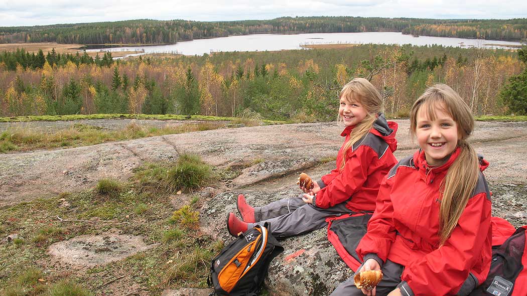 På berget sitter två leende barn med bröd i händerna. I bakgrunden öppnar sig ett höstlikt skogs- och sjölandskap.