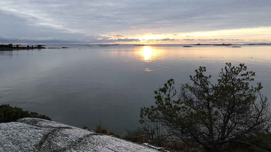 Havet och öar längre bort. På en mest molnig himmel skiner solen lågt.