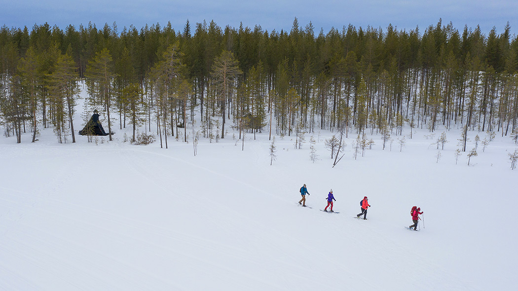 Fyra snöskor i flygvyn går från en timmerstuga över en snöig damm.