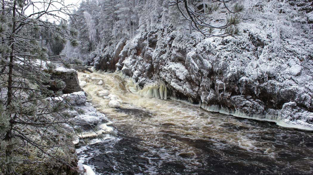 Röda klippväggar med ett tunt lager snö ovanpå. Mellan klipporna dånar en frostig fors.