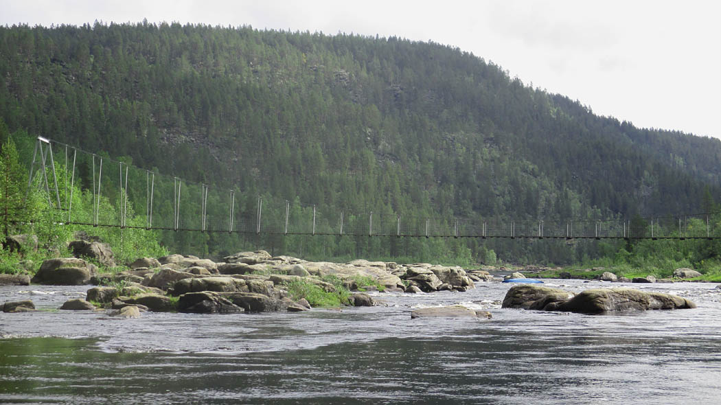 En snabbt strömmande flod med steniga stränder och stigande land på båda sidor. En smal hängbro av stål korsar floden.