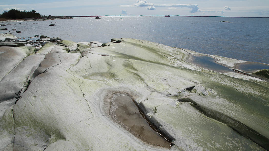 En slät strandklippa, i bakgrunden öppet vatten och på himlen lätta sommarmoln.