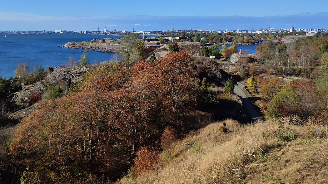 Höstlig landskap från en hög gräsbevuxen sluttning, i förgrunden träd, buskar och berg, samt en smal väg. I bakgrunden havet, stadens höga hus, torn och skorstenar.