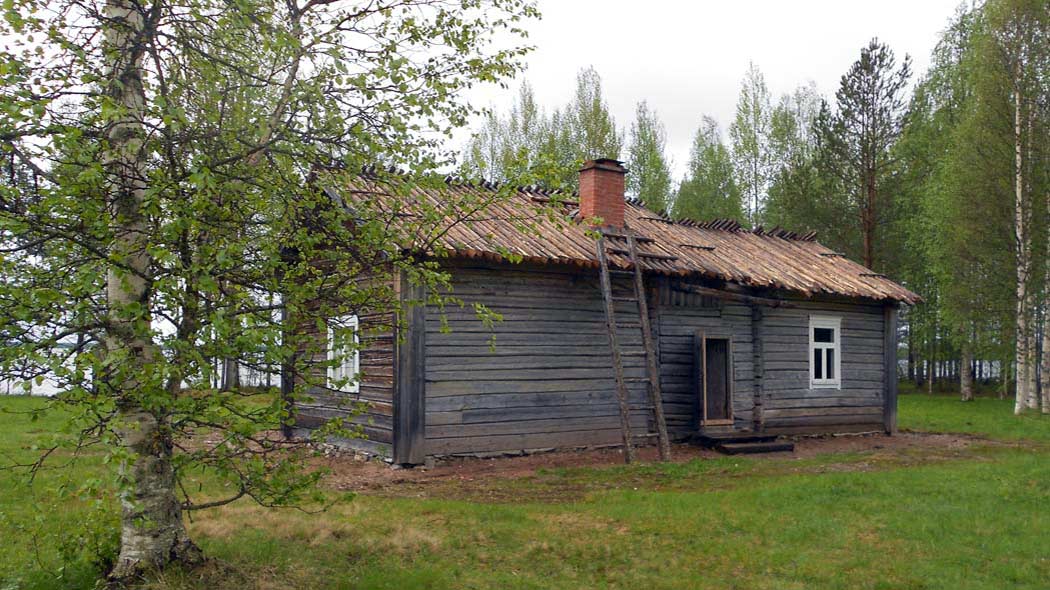 En grå byggnad mitt på gårdstunet. I bakgrunden syns träd och mellan träden skymtar en sjö. En björk med gröna löv finns till vänster i bild.