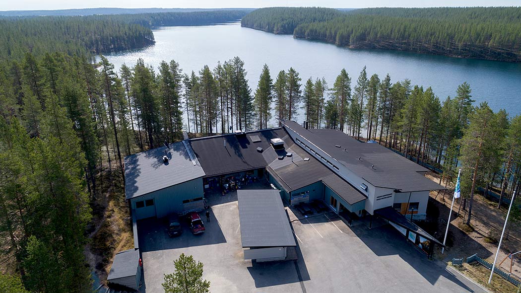 Ett flygfotografi av en byggnad, framför byggnaden finns bilar parkerade på en asfalterad gård. Skog omber byggnaden.