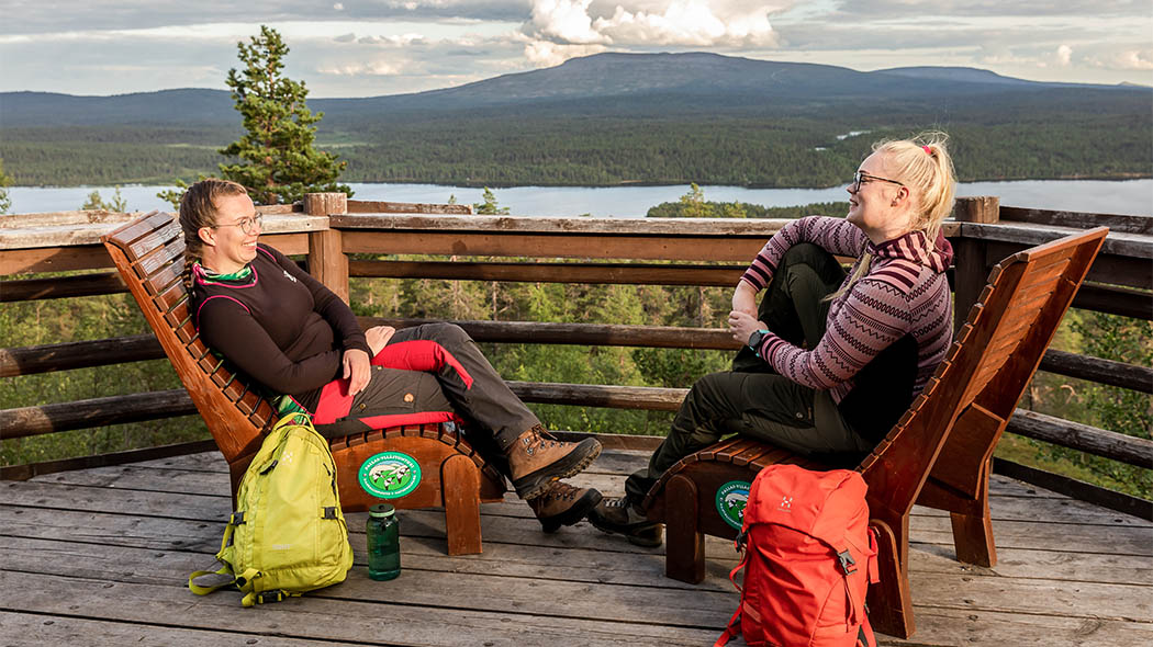 Två leende kvinnor sitter mitt emot varandra i trästolar bredvid ett räcke på utsiktsterrassen. I bakgrunden kan man se sommarlandskapet till fjället. Ryggsäckarna för vandrare ligger på marken bredvid stolarna.