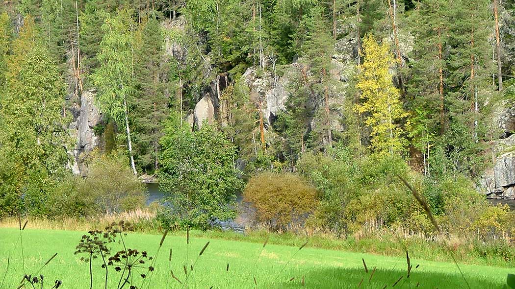 En somrig åker. I bakgrunden syns en sjö och branta klippor, tallar och granar växer på klipporna.