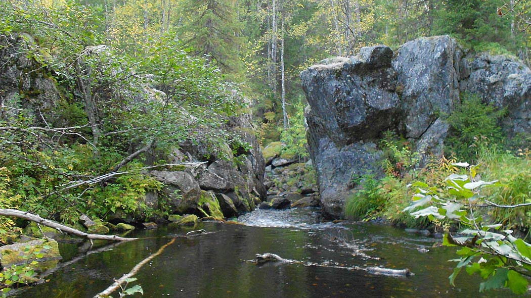 Mellan två stora stenblock strömmar en älv. I omgivningen finns grönskande växtlighet och skog.