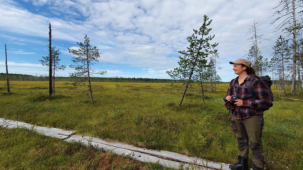 En glad person står högt och tittar på landskapet med en kamera i handen. I bakgrunden finns ett stort träsk.